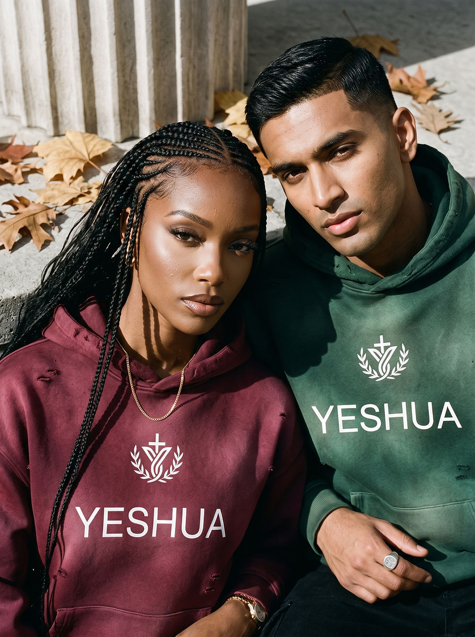 A female and male model sitting close together outdoors wearing matching distressed YESHUA hoodies in maroon and forest green.