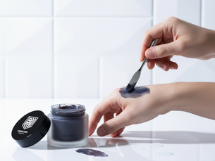 Hands applying dark cream from a jar labeled "Glow Mask" with a spatula on a white tiled background. Calm and clean atmosphere.