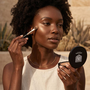 Woman outdoors applies makeup with a brush while holding a Yeshua Skin Cosmetics compact in warm sunlight.