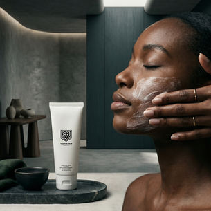 Woman applies face cream in a calm gray spa bathroom beside a Yeshua Skin Cosmetics tube on a tray.