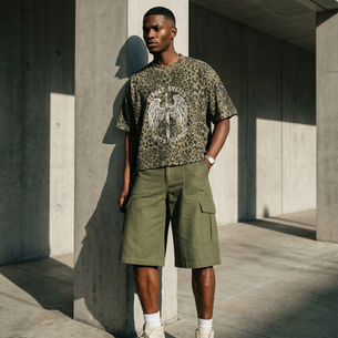 Man in leopard print shirt and green shorts leans against a concrete wall. Sunlit, neutral tones. Text on shirt: "Street Angels Gritty Soldiers."