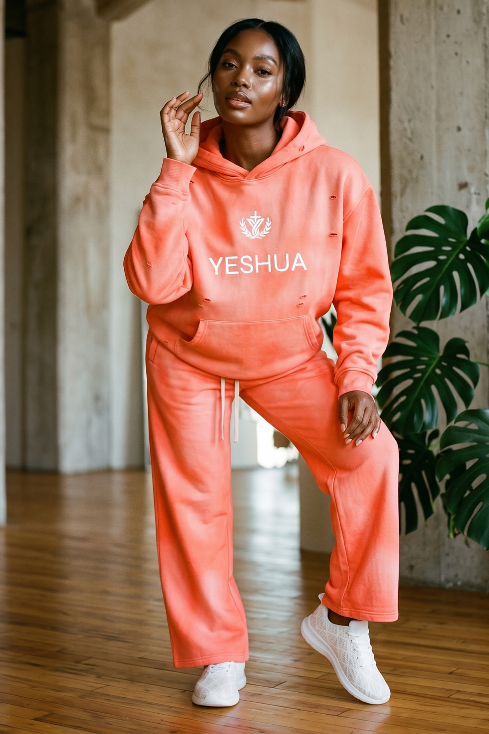 Woman in coral tracksuit with YESHUA text poses indoors beside a large green plant in a loft-like room.