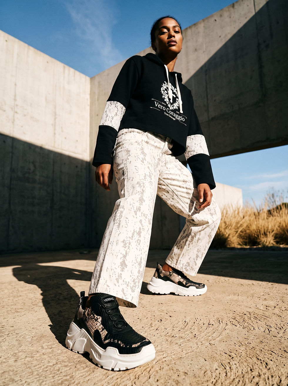 Low-angle outdoor shot of a female model posing near concrete walls under a blue sky, wearing a black "Vero Coraggio" cropped hoodie with lace sleeve panels, patterned off-white jeans, and chunky black and snakeskin platform sneakers.