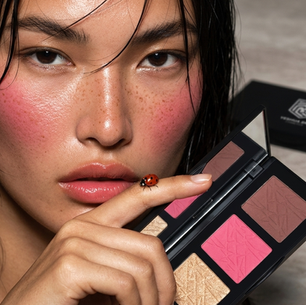 Close-up of a person with pink blush, holding a makeup palette. A ladybug sits on their finger. Neutral background, focused expression.