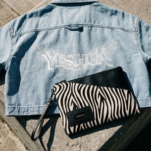 Denim jacket with "YESHUA" and wings, laid on concrete. Zebra-patterned clutch with "YESHUA" text beside it. Sunny setting.