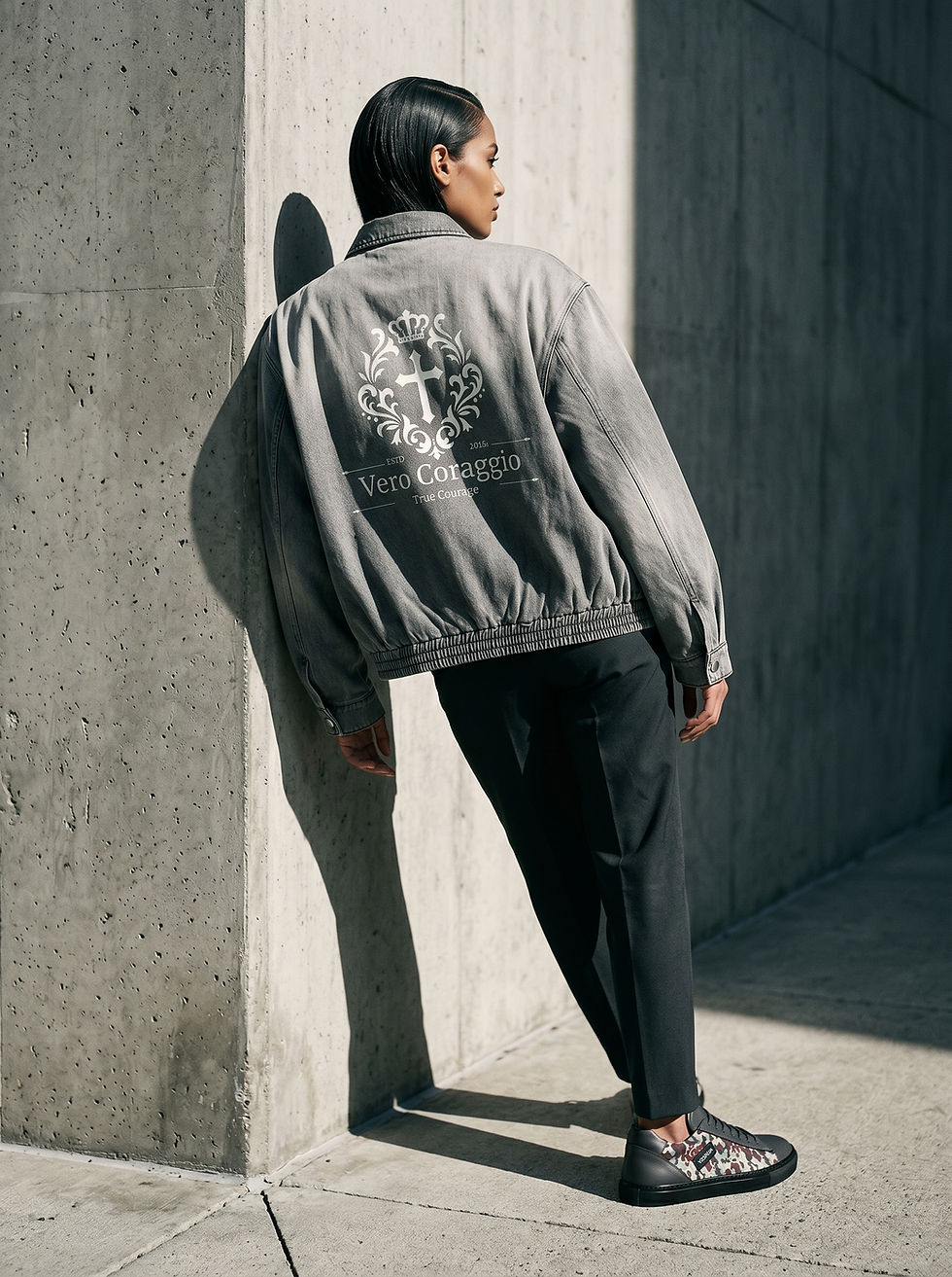 A full-length outdoor shot from behind of a person leaning against a concrete wall in the sunlight. They are wearing a gray wash denim jacket featuring a large white graphic crest and the text "Vero Coraggio True Courage" on the back, paired with slim-fit black trousers and camouflage-patterned sneakers.