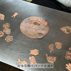 背景を彫金し終わった大仏の描かれた銅板。取り付ける錺金具のパーツ（猫や亀など）が散らばっている