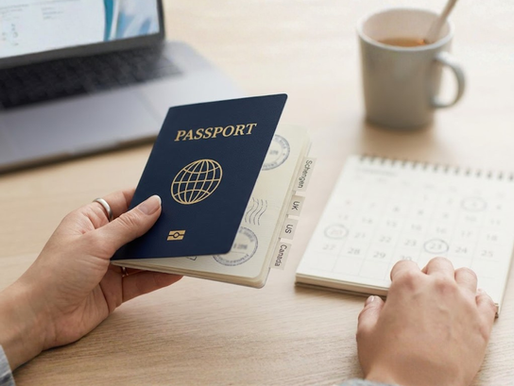 Close-up of a person holding a generic navy blue passport with bookmark tabs labeled Schengen, UK, US, and Canada, resting on a wooden desk next to a blurred calendar.