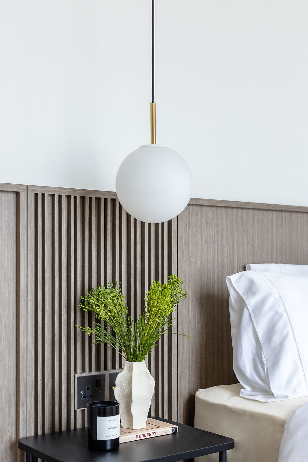 Modern bedroom nightstand with white pendant lamp, vase of green flowers, BYREDO WOODS candle, and BURBERRY book near white bedding