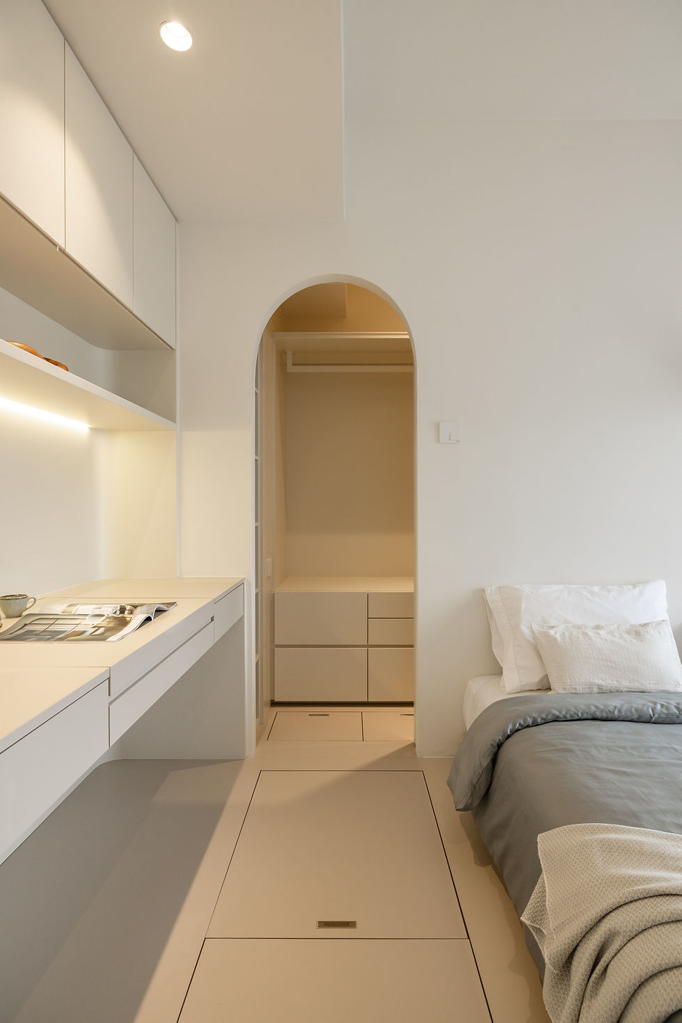Minimalist cream bedroom with bed, built-in desk, and arched closet doorway, softly lit and calm