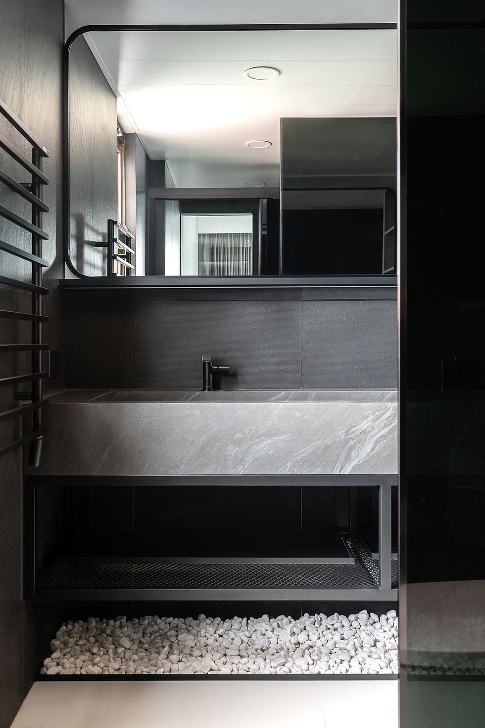 Sleek modern black bathroom with large mirror, dark vanity, stone sink, and white pebbles below in a minimalist spa-like setting