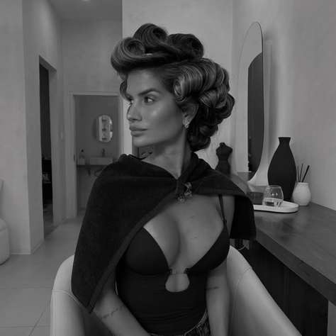 Woman with roller-set hair and dark shawl sits in salon, neutral expression. Minimalist decor with a mirror, vases, and soft lighting.