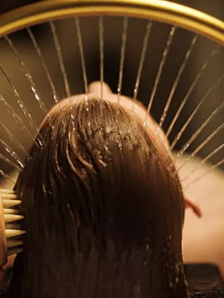 Person lying under a golden arc fountain with hair being brushed. Water streams evenly, creating a relaxing spa atmosphere.