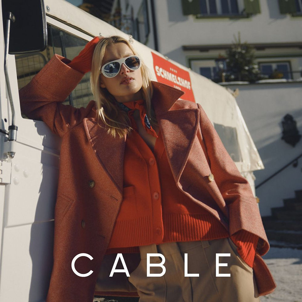Blonde woman in orange coat leans on a white vehicle. She's wearing sunglasses. Background shows steps and buildings. Text reads: CABLE.