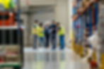 Five people in a warehouse, wearing yellow vests, discuss papers. Shelves with goods in the background. Professional, focused atmosphere.