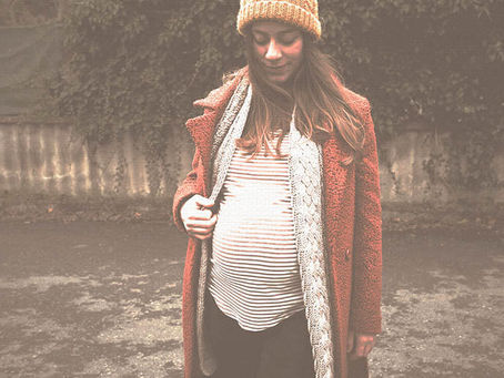 Pregnant woman in an orange coat and knit hat stands outdoors, looking down against a weathered wall and pavement.