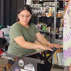 Woman in green shirt sits at glassblower's or gaffer's bench rolling the blow pipe to keep the glass centered on the pipe, allowing the hot glass to cool.