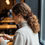 Thumbnail: A woman sits in a cafe. Her curly hair is tied in a sophisticated ponytail held with a cocoa coloured silk scrunchie