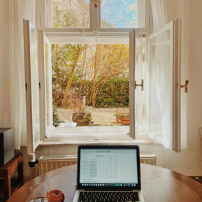 Open laptop on round table with spreadsheet, next to a glass of water and an apple. Bright open window shows garden under clear blue sky.
