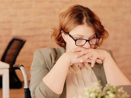 Woman with glasses in a blazer sits at a desk, hands clasped, appearing thoughtful. Background features a brick wall and office setting.