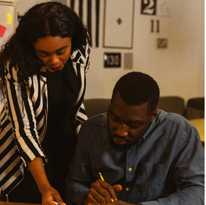 A woman in a black and white striped blazer leans over a table, pointing at documents while a man in a blue denim shirt writes with a pencil. Both are focused on the work in front of them in a warmly lit office space with graphic art on the wall in the background.