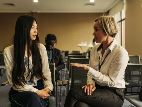 Two women in conference chairs engage in conversation, wearing lanyards. A third person sits behind them. The setting is a well-lit room.