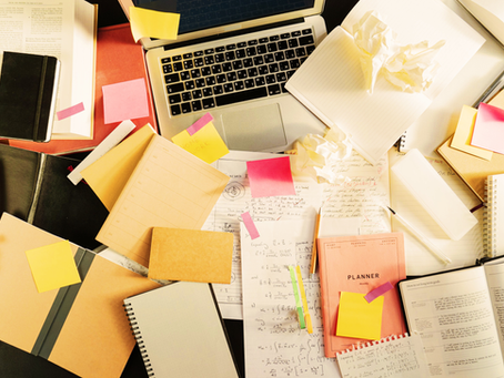 An overhead view of a chaotic desk covered in open notebooks, journals, and loose papers filled with handwritten notes and mathematical equations. Sticky notes in yellow, pink, and orange are scattered throughout. A laptop sits open near the top of the frame surrounded by the chaos. A monthly planner, crumpled paper, pencils, and highlighters are visible among the clutter. Every inch of the surface is covered.