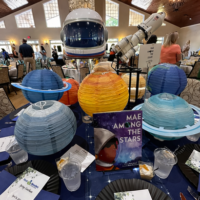 Outer space themed table featuring ‘Mae Among the Stars’ book, astronaut helmet, and planet decorations at Ferst Readers Henry County Spring Luncheon event
