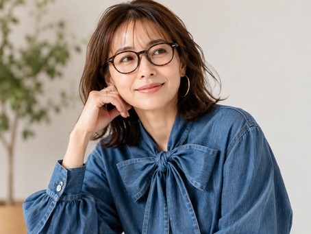 眼鏡をかけた40代女性顔タイプアクティブキュートが青いデニムのボウタイブラウスとフープピアス、伊達メガネを着て微笑み、手を頬に添えて室内でくつろぐ。背景はぼけた観葉植物。