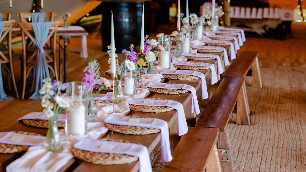 Long rustic banquet table set with flowers, candles, woven placemats, and lavender napkins in a warm tented event space