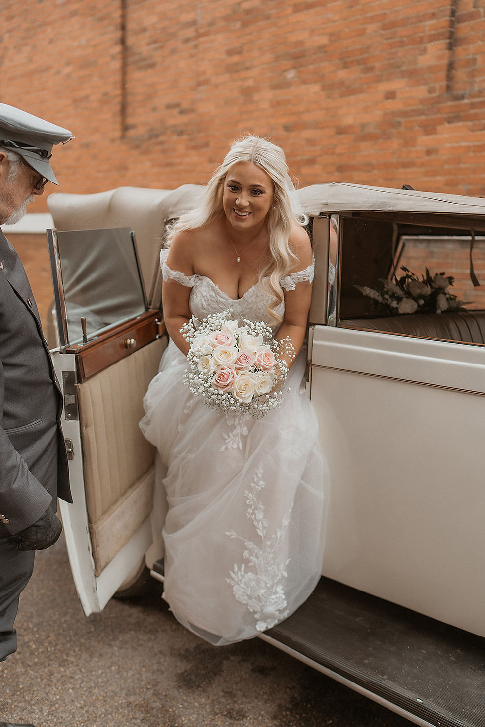 Bride in white dress holding pink and white bouquet, exiting vintage car, brick wall background, smiling at chauffeur.