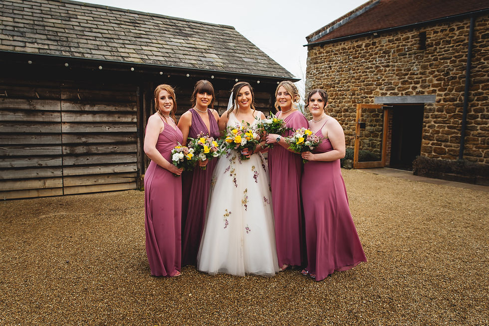 Bridal party with bride in white gown and four bridesmaids in mauve dresses holding bouquets, standing outdoors by rustic wooden buildings.