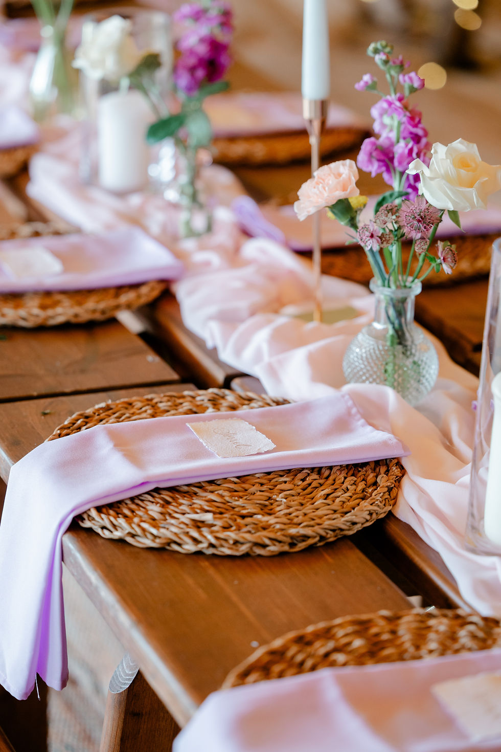 Wedding table settings with lavender napkins, woven placemats, and small flower vases; a tag reads Bride.