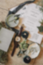 Wedding invites with greenery theme, text for Chelsea and Aidan. Nearby are a watch, ring, and jewelry on a wooden table. Calm, elegant mood.