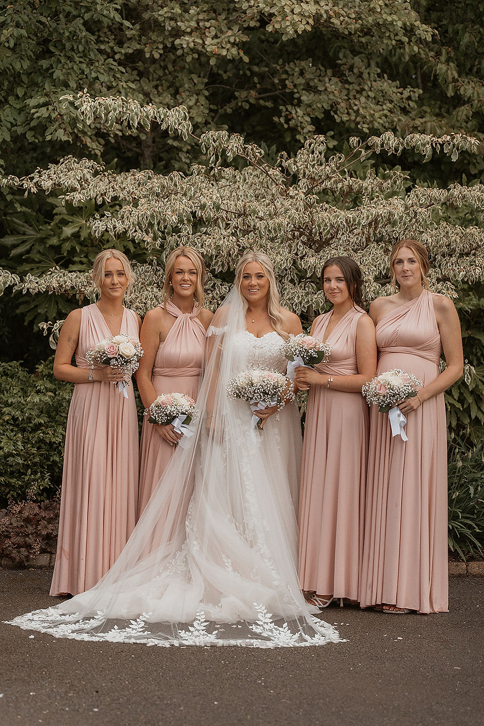 Five women in pink dresses and one in a white wedding gown stand smiling, holding bouquets, with lush greenery in the background.