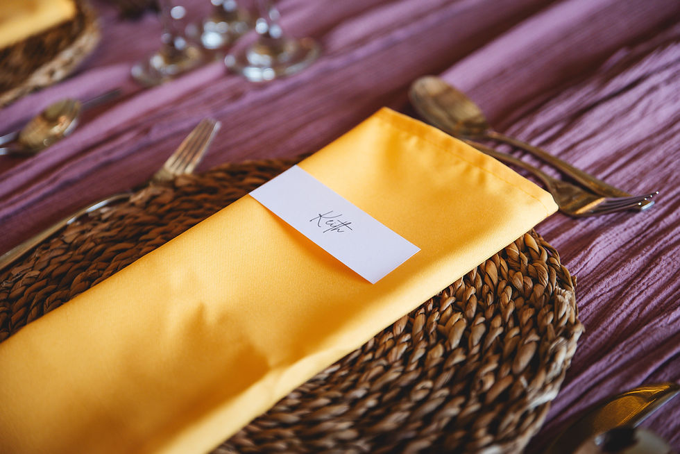 Yellow napkin with name card "Keith" on woven placemat, surrounded by silver cutlery on a purple tablecloth. Elegant dining setup.