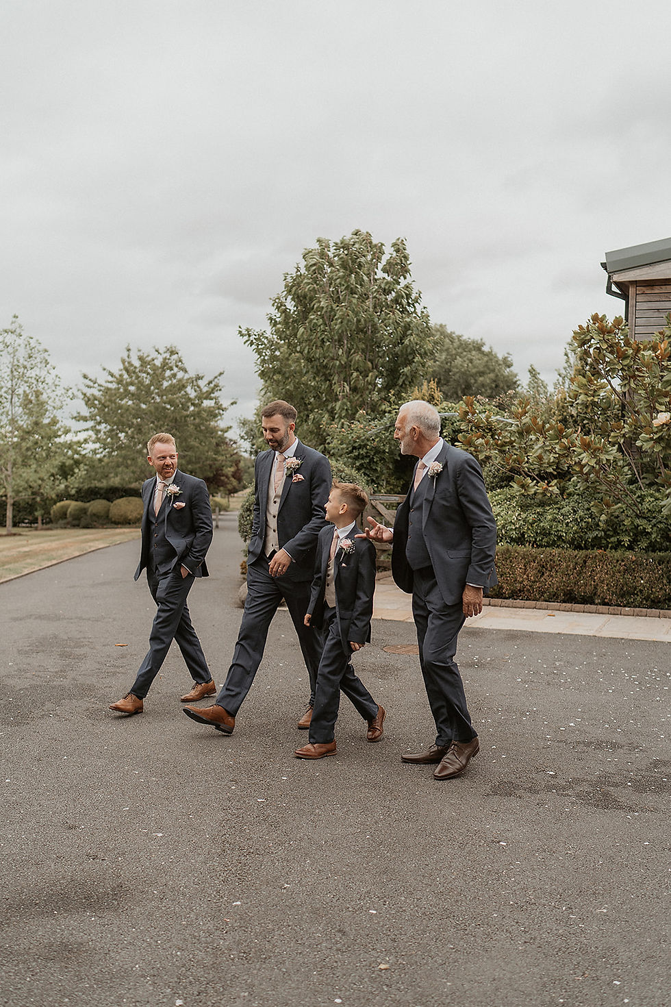 Four men in suits, including a child, walk on a paved path in a garden setting. Overcast sky; greenery and a building in the background.