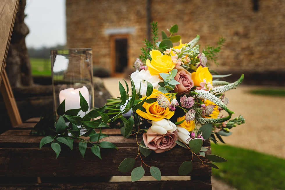 Bouquet of yellow and pink roses with greenery on a wooden table. A lit candle in a glass holder nearby. Stone building and greenery backdrop.