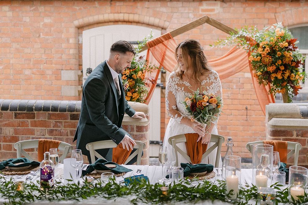 Bride holding flowers and groom adjust chairs at a decorated outdoor table. Orange and green accents, brick wall, cheerful atmosphere.