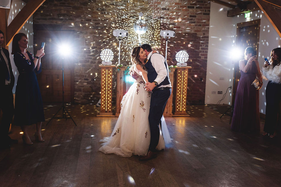 Couple dancing at a wedding in a rustic hall, surrounded by guests taking photos. Disco lights and warm ambiance create a festive mood.