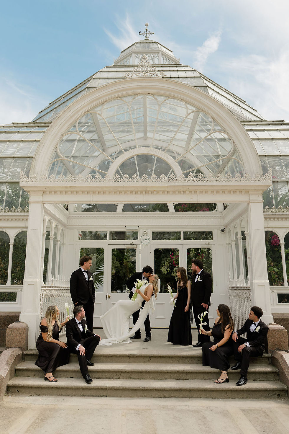Wedding party poses outside a glass conservatory; groom dips and kisses bride while bridesmaids and groomsmen in black watch.