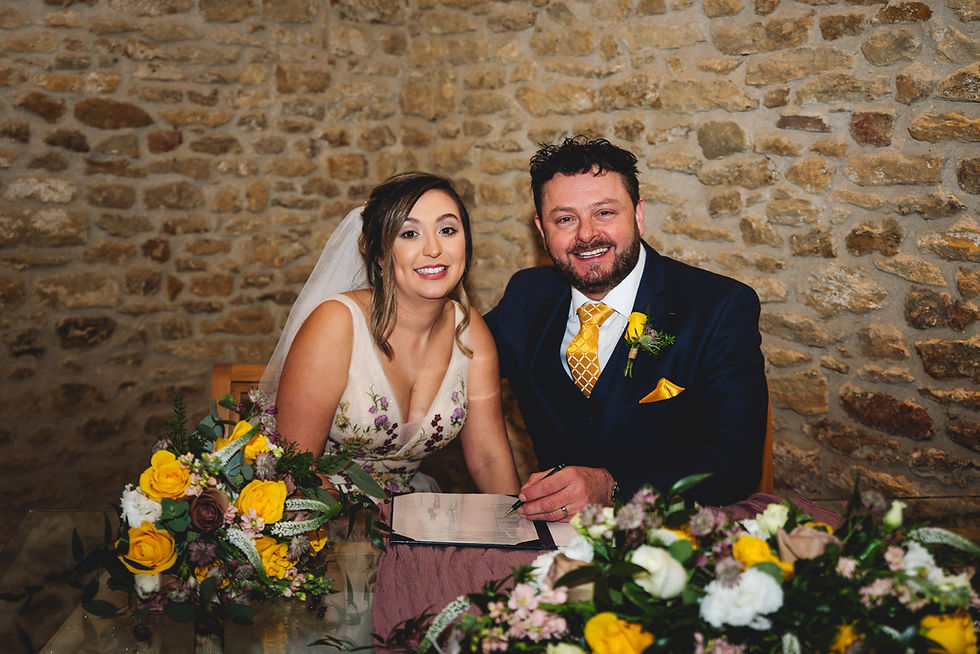Bride and groom smiling, signing a document in a stone-walled room. Bright floral bouquets with yellow accents surround them.