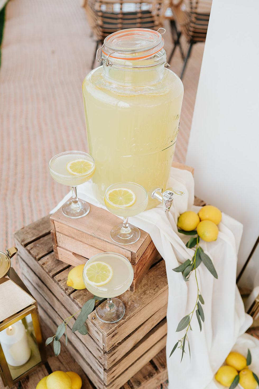 Lemonade in a large glass container with tap, surrounded by lemons and leafy branches. Three filled glasses on wooden crates, outdoor setting.