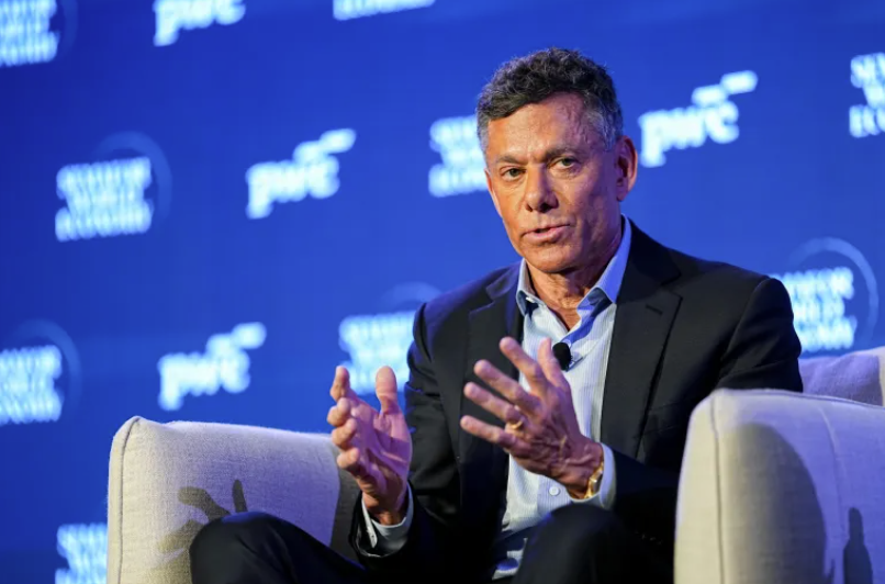 Strauss Zelnick addresses an audience during a conference, gesturing as he discusses key insights on a stage with a blue backdrop featuring event sponsors.
