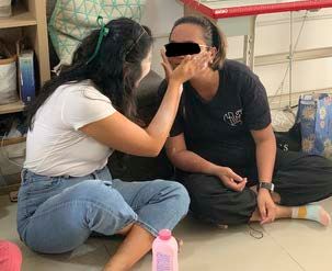 Two women sitting on the floor during a beauty training session, one gently applying lotion to the other’s face with kindness and care. (Face partially obscured for privacy.)