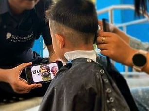 Young boy receiving a haircut from ShearLove students during an outreach event, with another team member showing him a video on a smartphone for comfort.