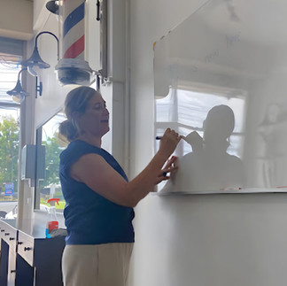 A woman teaching at Shear Love International in Thailand, writing on a whiteboard inside a classroom with a barber pole visible through the window, reflecting the vocational training setting.
