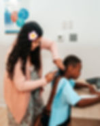 Woman in pink cardigan examines a boy in a blue gown in a clinic. She uses a thermometer on his ear. Poster and thermostat in background.