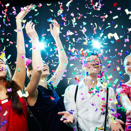 Four people celebrating, smiling joyfully under colorful confetti. They're dressed in festive attire with party hats, against a vibrant, lit backdrop.