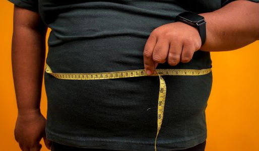 A close-up of a person measuring their waist circumference with a yellow tape measure over a dark t-shirt, against an orange background. The image illustrates abdominal weight gain as one of the key indicators of metabolic health risk.